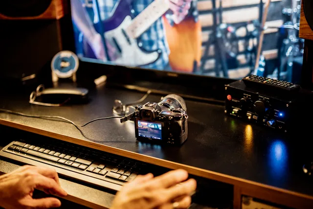 A person sits at a keyboard in a music studio. A camera is placed on a desk, connected to a computer screen showing musicians. Sound equipment is visible nearby. Creator studio equipment UK regulations by activity